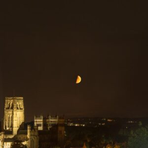 In this nighttime photograph, the majestic silhouette of Durham Cathedral looms powerfully against a deep, inky sky. The cathedral's ancient Norman architecture is awash in a warm, golden hue, its robust towers and intricate stonework glowing with a timeless grandeur that echoes a millennium of history. Above, a crescent moon hovers low, casting a gentle, otherworldly light that contrasts beautifully with the darkened expanse. The distant lights of the city flicker softly on the horizon, blending the medieval with the modern in a subtle, harmonious dance. The interplay of light and shadow crafts a hauntingly beautiful scene, where the enduring spirit of the past meets the quiet stillness of the present under the moon's watchful glow.