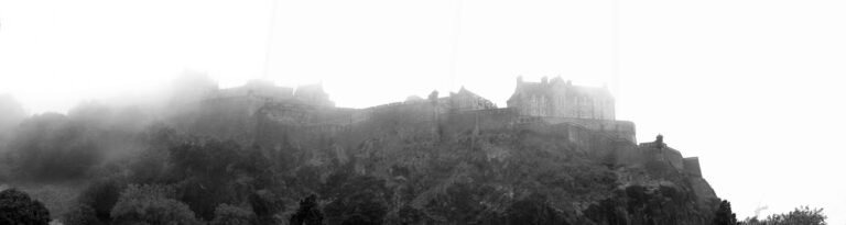Dramatic monochrome panorama of Edinburgh Castle shrouded in thick morning mist on its rocky crag.