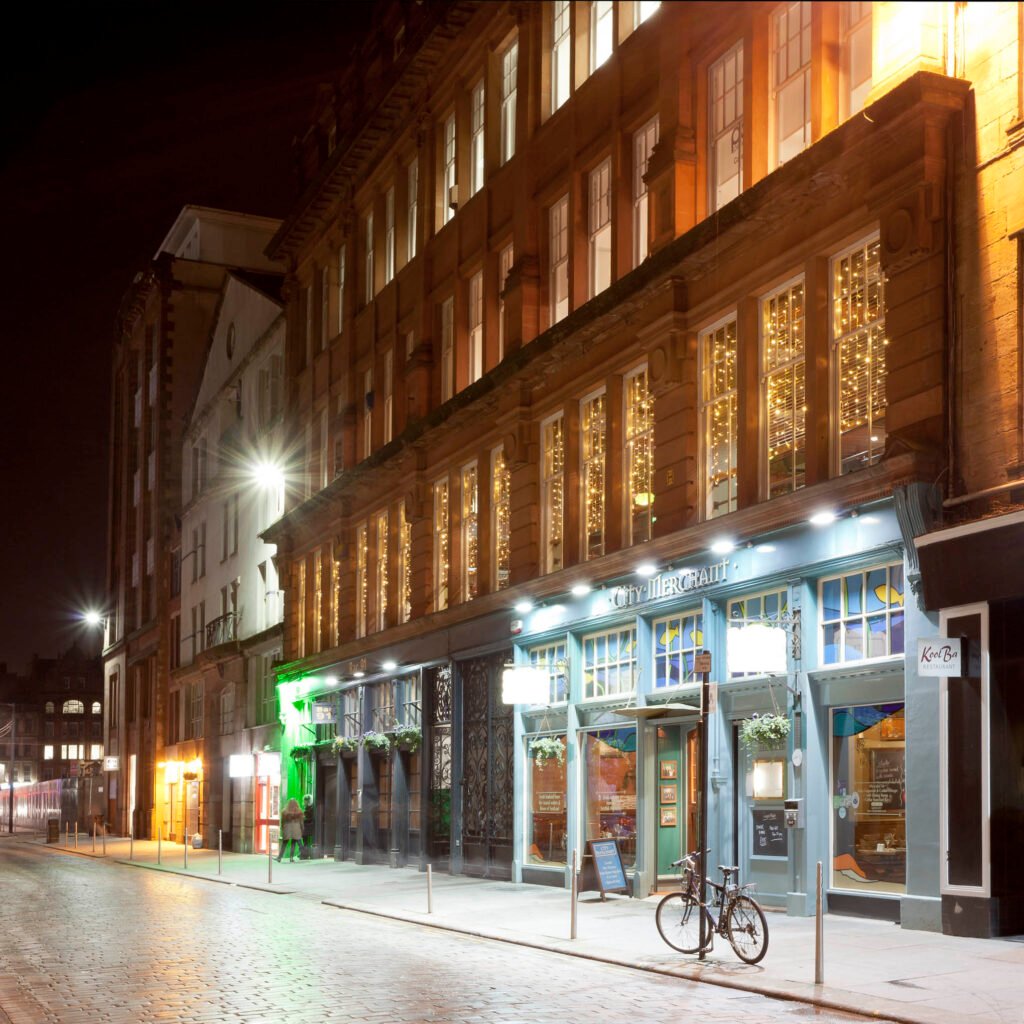 Night photograph looking down a cobbled street in Glasgow past illuminated red sandstone buildings. Cold blue LED downlights highlight a restaurant storefront with a bicycle parked outside, contrasting with warm amber lights and fairy lights glowing in the upper-floor windows. Scotland. Author : Stephane Loustalot,