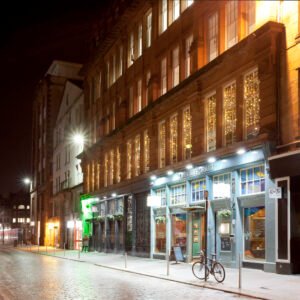 Night photograph looking down a cobbled street in Glasgow past illuminated red sandstone buildings. Cold blue LED downlights highlight a restaurant storefront with a bicycle parked outside, contrasting with warm amber lights and fairy lights glowing in the upper-floor windows. Scotland. Author : Stephane Loustalot,