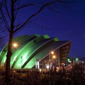 Night photograph of Glasgow. When looking at modern architecture, it is interesting to look at the surroundings and see how nature is added. In this photograph, although we see that the edge has been trimmed, the overall effect is rather wild. In this island of nature we also have a tree. It is not a lone tree as we can see the branches of second one out of the frame. Next to the imposing building it looks frail and unassuming, out of place. Photographer : Stephane Loustalot.