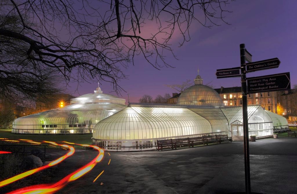 Long exposure photograph of the illuminated Kibble Palace glasshouse in Glasgow at twilight, featuring sweeping red and yellow vehicle light trails in the foreground, a dark purple sky overhead, bare winter tree branches framing the top, and a directional park signpost on the right.