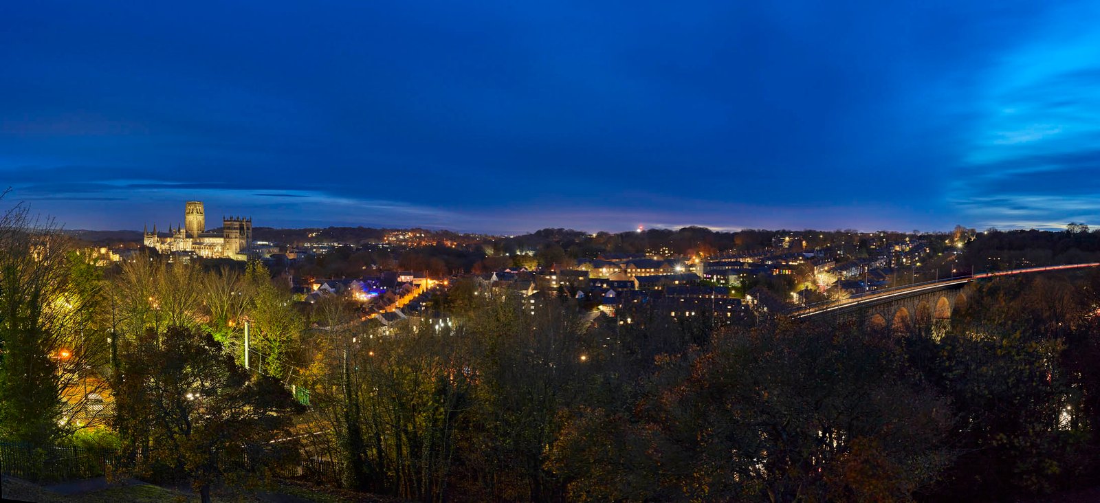 Panoramic night view of Durham featuring the illuminated Durham Cathedral on the left and the city viaduct glowing against a deep blue twilight sky.